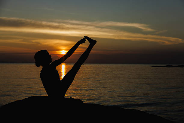 A silhouetted female performing a yoga pose on the seaside at sunset. The calm sea and warm colors create a serene and peaceful ambiance, evoking relaxation, mindfulness, and inner balance.