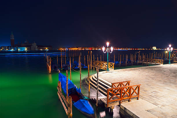 Romantic night view of Venice with gondolas moored along illuminated waterfront. Glowing street lamps and calm waters create a serene atmosphere in this iconic Italian cityscape