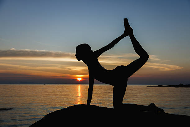 A silhouette of a woman performing a yoga pose against the backdrop of a serene ocean and vibrant sunset, highlighting mindfulness, fitness, and tranquility.
