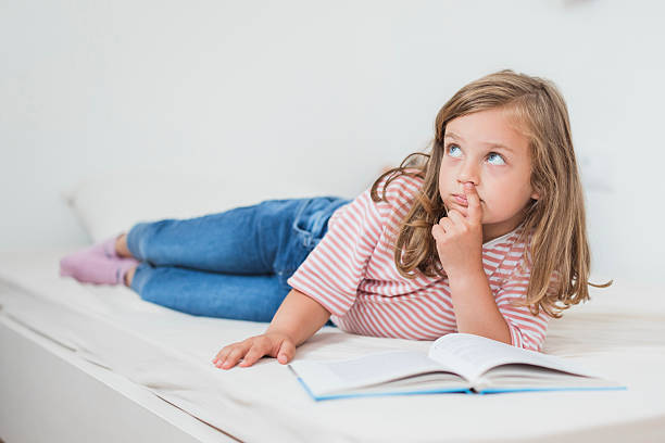 Elementary age girl lying on bed, reading a book and thinking with finger on nose