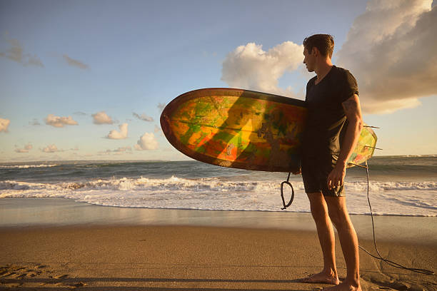 A surfer holds a colorful surfboard on a beach as the ocean waves crash ashore and sunlight bathes the scene, evoking themes of adventure, relaxation, and connection with nature.