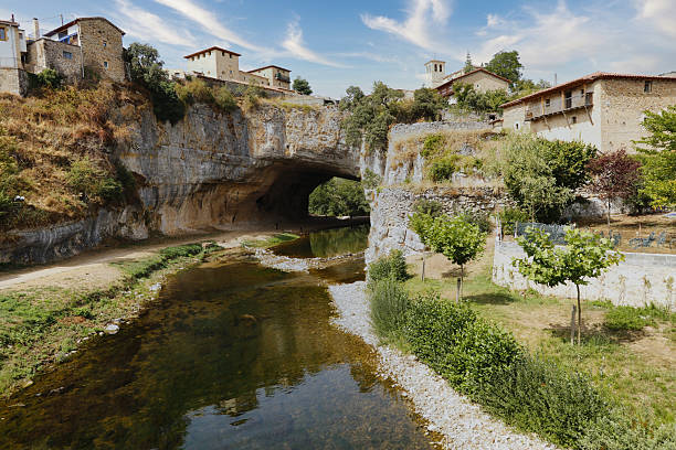 istock Natural rock arch over a serene river - A river flows through a natural rock arch, with ancient houses perched on the cliff above under a blue sky. 2234896322