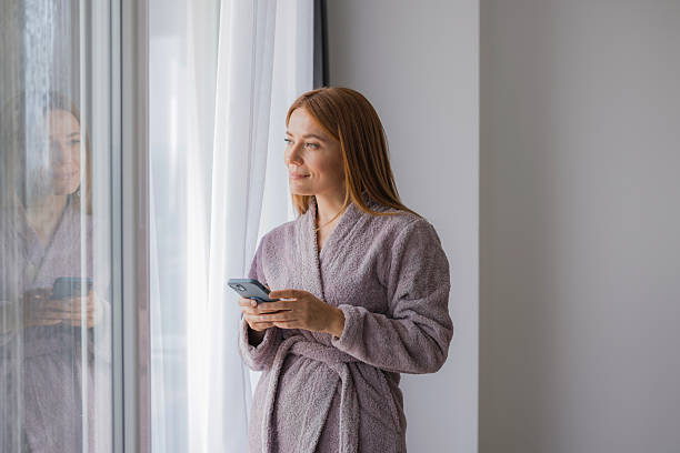 istock Smiling woman wearing bathrobe using smartphone by window 2234897324