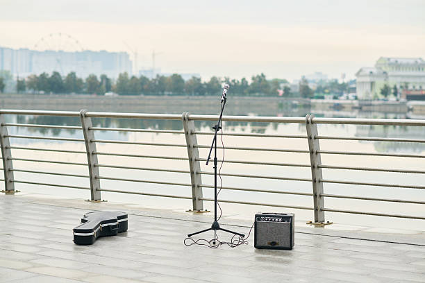 Empty riverside promenade featuring microphone on stand, guitar amplifier, and closed guitar case positioned near metal railing, cityscape and ferris wheel visible in background