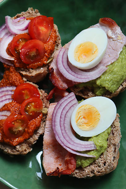 Full frame image of green plate of open sandwiches, halved homemade multigrain bread rolls with pumpkin and sunflower seeds, toppings of guacamole, bacon rashers, red onion and boiled egg halves or red pesto sauce, red onion and cherry tomato halves