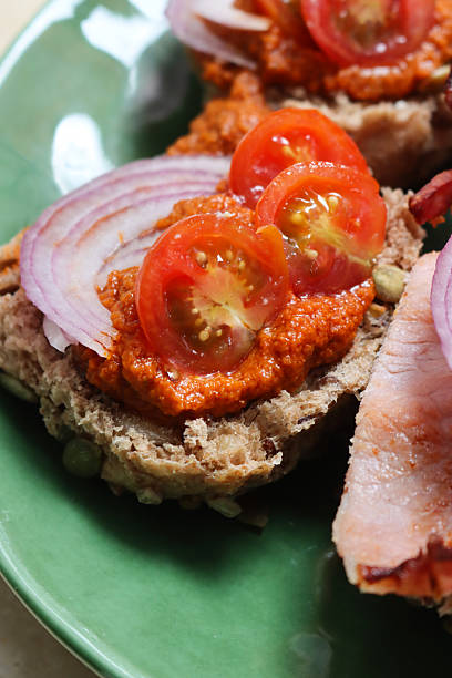 Full frame image of green plate of open sandwiches, halved homemade multigrain bread rolls with pumpkin and sunflower seeds, toppings of guacamole, bacon rashers, red onion and boiled egg halves or red pesto sauce, red onion and cherry tomato halves