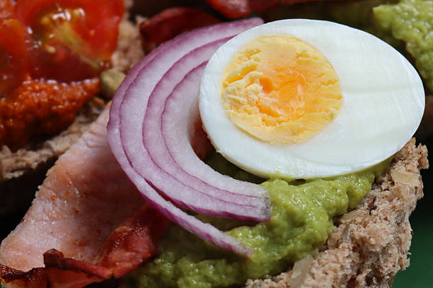 Stock photo showing close-up, elevated view of open sandwiches made from homemade, multigrain bread roll halves topped with either guacamole, bacon rashers, red onion slices and hard-boiled egg halves, or red pesto sauce, sliced red onion and halved cherry tomato.