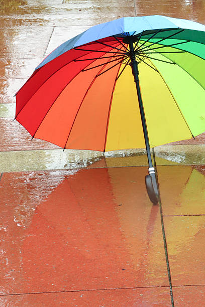 Stock photo showing close-up view of a rainbow umbrella left on a paved area. The wet monsoon weather creates puddles and reflective surfaces during the heavy rain.