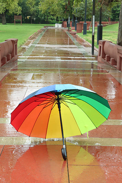 Stock photo showing close-up view of a rainbow umbrella left on a paved area. The wet monsoon weather creates puddles and reflective surfaces during the heavy rain.