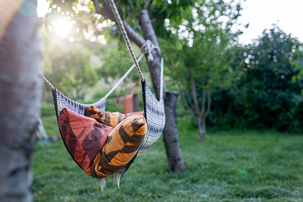 Photo of empty home made hammock in the garden. Green grasses and green trees seen around. No people are seen in frame. Shot with a full frame mirrorless camera.