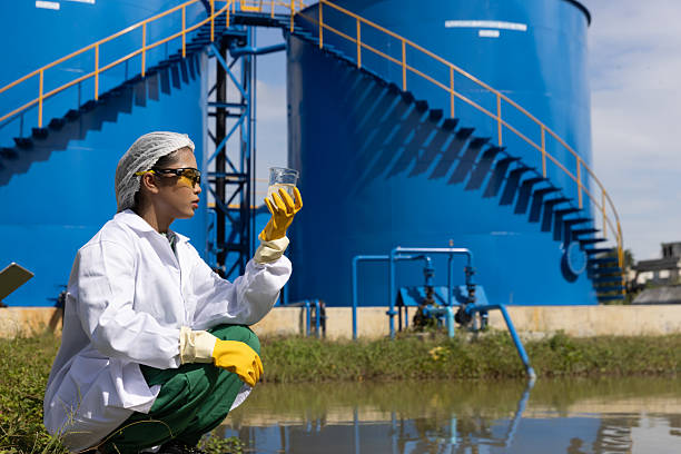 Young science Biologist or environmental quality inspector wearing protective gear and white lab coat collecting water sample from Sedimentation pond , water treatment facilities