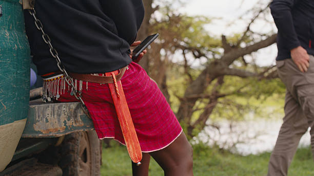 A Maasai man wearing a traditional shuka using a smartphone while leaning against a vehicle in rural Kenya. The scene features natural surroundings with trees and open landscape, highlighting cultural and technological blend.