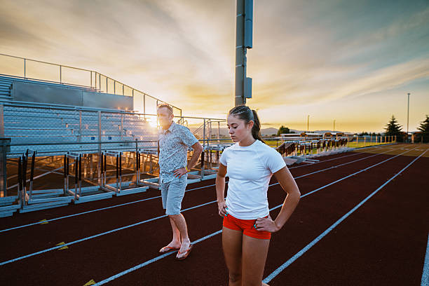 A female collegiate track and field athlete walks with her coach on the school stadium track at sunset, pausing to review performance after a one-on-one training session.
