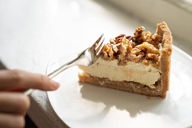 A slice of cheesecake topped with nuts served on a white plate by the window, with a hand holding a fork ready to take a bite.