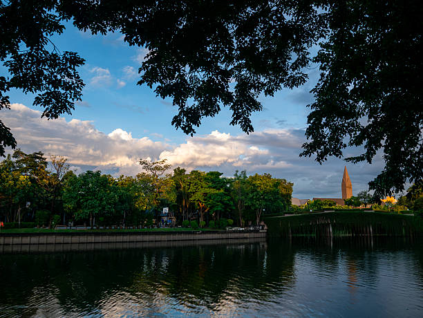 istock Serene evening waterfront park scene with lush green trees reflecting in calm river under dramatic cloudy sky during golden hour. 2234957132