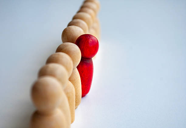 Side view of beige wooden dolls aligned in a line with one red painted doll among them highlighting individuality or difference