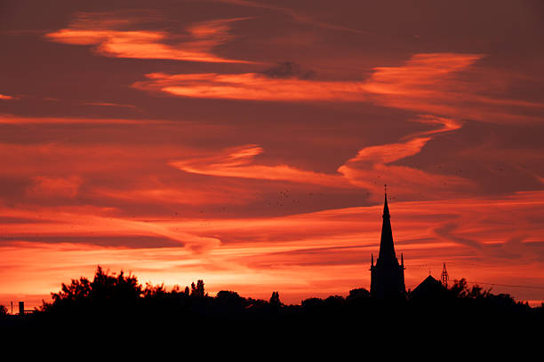 A breathtaking sunset with deep red clouds and the dark silhouette of a church in the background, while birds fly across the colorful evening sky. Atmospheric landscape photography capturing mood and contrast.