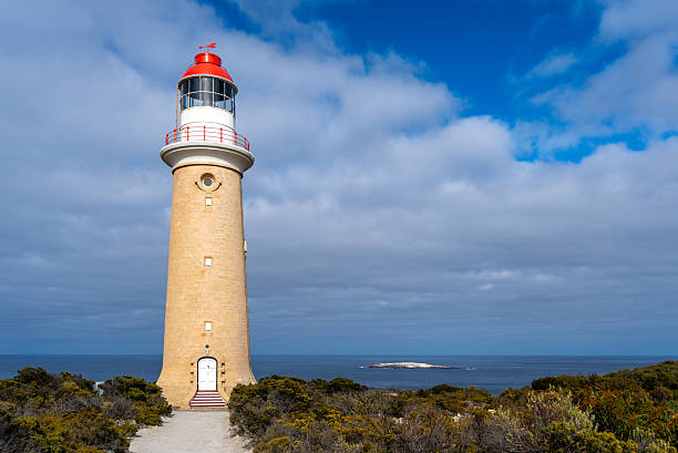 The Cape Du Couedic lighthouse stands tall amidst coastal vegetation in Flinders Chase National Park, Kangaroo Island, Australia. Its red and white top contrasts with the cloudy sky
