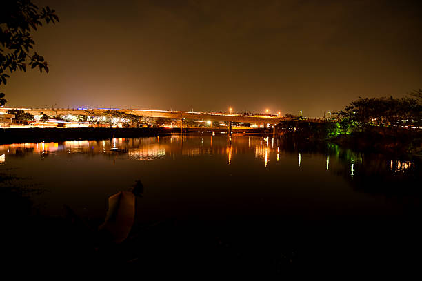 Nighttime urban scene featuring illuminated city structures and bright lights reflected on a tranquil river.