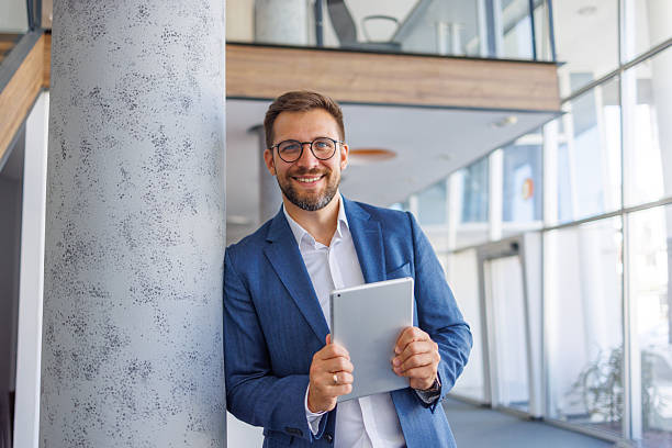 istock Smiling businessman leaning against pillar holding digital tablet in modern office 2234972011