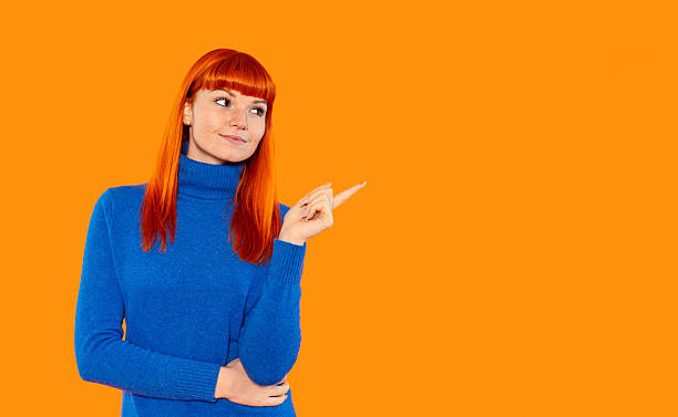 istock Woman with vibrant orange hair points confidently against a bright orange background while wearing a blue turtleneck sweater indoors during daylight hours 2234976120