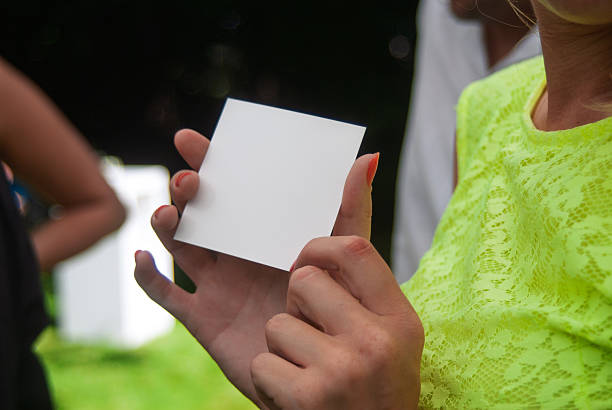 A close-up view of a person holding a blank square card, photographed in an outdoor setting with greenery as a background. The vivid colors add contrast and draw attention to the card.