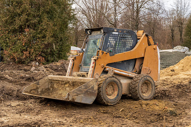 istock Orange skid steer loader parked on muddy ground. Heavy construction equipment with dirty bucket on a site with dirt piles and trees. 2234995306