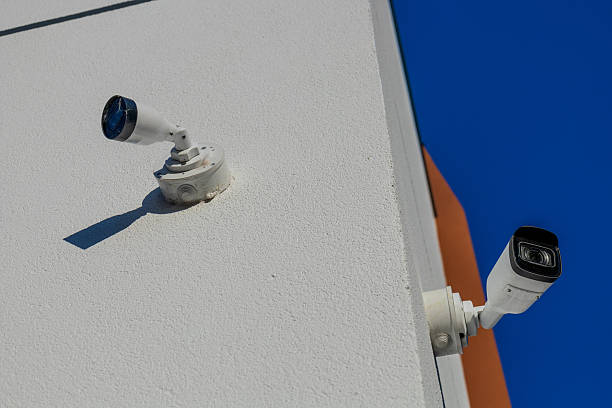istock White security camera and outdoor light fixture mounted on a textured building wall. Clear blue sky, bright sun, long shadows. 2234995341