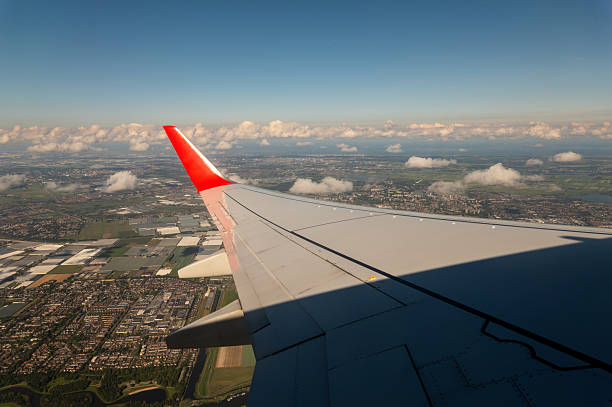 Airplane wing flying over cityscape and green fields, with white clouds and blue sky