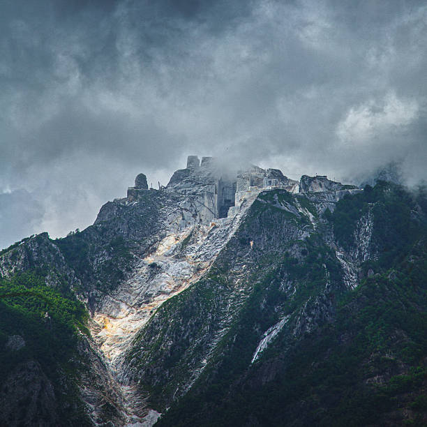 A dramatic marble quarry cuts bright white terraces into the dark mountainside of the Apuan Alps above Pietrasanta, with heavy clouds shrouding the peaks where centuries of extraction have carved geometric scars into the natural landscape.  This powerful industrial landscape symbolizes the intersection of human ambition and natural resources, representing both the creative potential of marble sculpture and the environmental impact of centuries-old extraction practices.