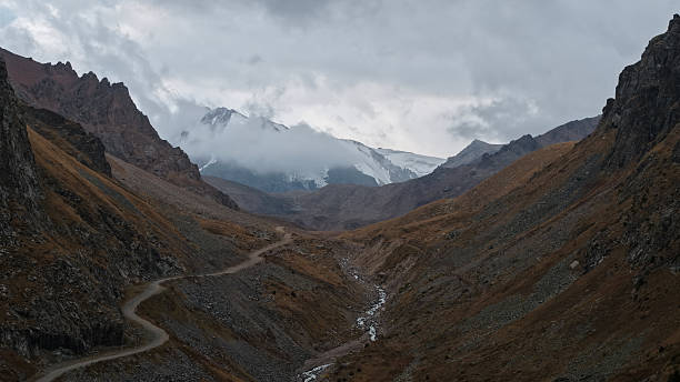 Majestic mountain valley features a winding dirt road leading through rocky terrain, surrounded by towering peaks and dramatic clouds, creating a breathtaking natural landscape