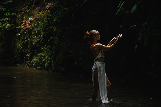 A serene image of a woman in a forested area, balancing elegantly in a gentle stream, surrounded by lush greenery and showcasing a connection with nature. The peaceful ambiance evokes tranquility and harmony.