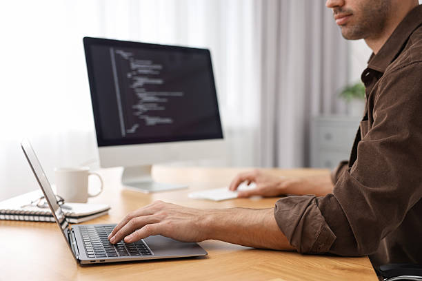 istock Programmer working on laptop and computer at wooden desk indoors, closeup 2235013718