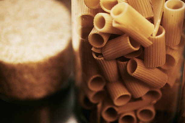 Close-up of golden rigatoni pasta inside a glass jar, paired with a rustic background featuring grains, creating a warm kitchen aesthetic.