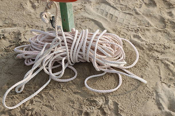 Close up of a white rope coiled on the sand, next to a metal pole, on a beach, suggesting maritime or rescue activities