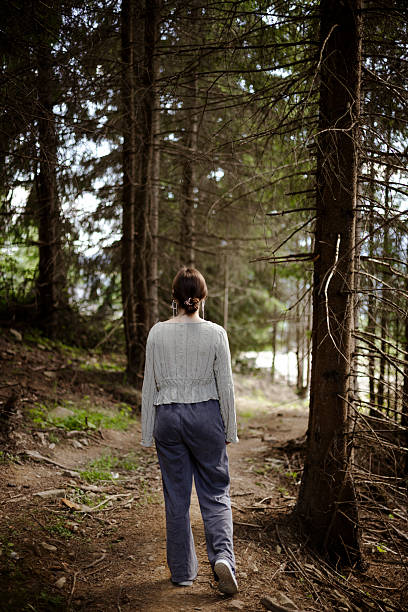 Back view of young woman walking alone in wild forest along path. Full length body lifestyle scene of freedom and solitude in nature.