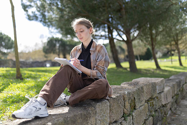 istock Young woman writing in notebook sitting on stone wall in park 2235031997