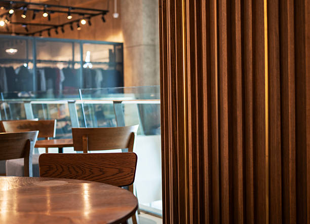 A warm cafe interior featuring a wooden slatted partition, round wooden tables, and seating areas. Track lighting and a glass barrier create a modern yet inviting dining atmosphere.