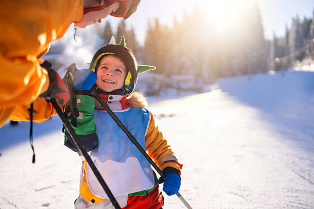 A young child, dressed warmly in ski gear, smiles enthusiastically while learning to ski in the mountains on a sunny winter day, guided by an attentive instructor under clear skies.