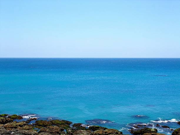 A tranquil ocean scene featuring crystal-clear blue waters stretching to the horizon beneath a cloudless sky. In the foreground, rocky formations partially submerged in shallow water reveal patches of green algae and moss, adding texture and depth. The gradient from light to deep blue evokes calm and clarity, making this image ideal for travel promotions, environmental campaigns, wellness branding, and minimalist design. It captures the quiet beauty of coastal nature and the soothing rhythm of the sea.