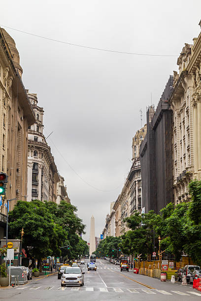 Buenos Aires, Argentina - March 14, 2020: Cars navigating the bustling Avenida 9 de Julio, heading towards the iconic Obelisk in Buenos Aires, Argentina, while a cloudy sky looms overhead, creating a dynamic cityscape