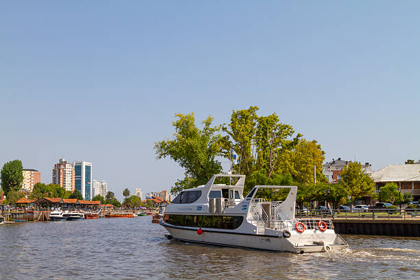 Buenos Aires, Argentina - March 13, 2020: White tourist boat navigating the Tigre River in Buenos Aires, Argentina, surrounded by moored vessels and modern buildings under a clear blue sky on a bright, sunny day