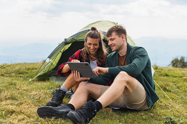 Modern young couple camping, enjoying time outside their tent, laughing, sharing moments, and streaming on a tablet in a stunning natural setting.