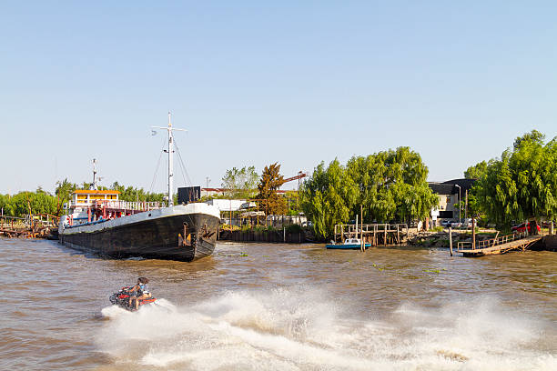 Young adult is riding a jet ski, creating waves and splashes, near a large cargo ship in the Riachuelo river, Buenos Aires, Argentina, on a sunny day, with trees and buildings on the shore