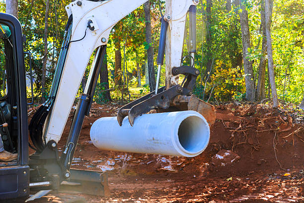 An excavator is in action, lifting large cement pipe amidst construction site in wooded area.