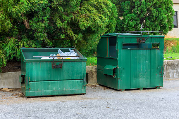 istock Two green dumpsters positioned beside concrete wall in parking area surrounded by trees on clear day 2235075155