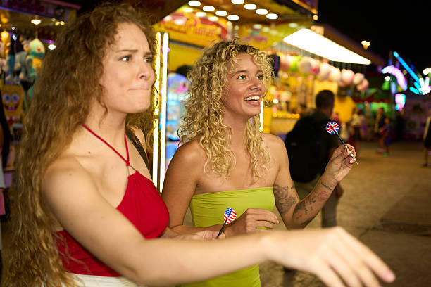 Two young women enjoying an evening at an amusement park, playing darts together and sharing smiles under colorful lights