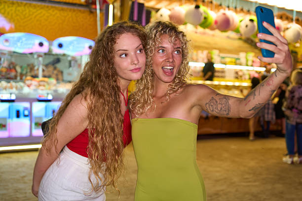 Two friends capturing a joyful selfie at the amusement park, reveling in the excitement of their evening adventure filled with colorful lights