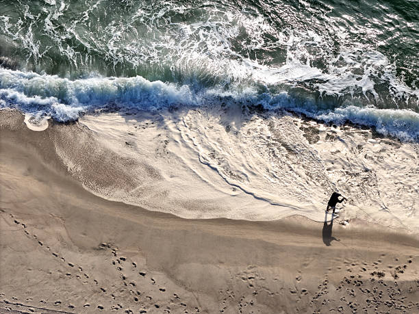 Aerial view of waves crashing against sand beach as fisherman fishes along shore.