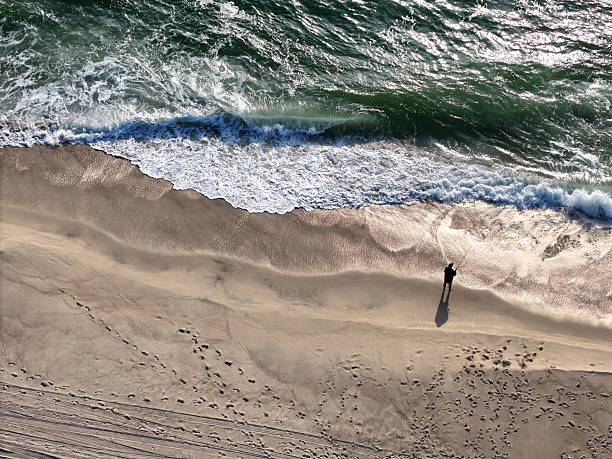 Aerial view of waves crashing against sand beach as fisherman fishes along shore.
